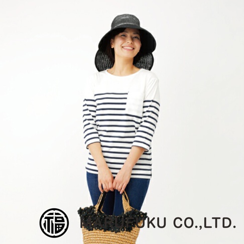 一位面帶微笑的女性模特兒，頭戴黑色抗UV遮陽寬緣帽，身穿白色底搭配海軍藍橫條紋的三分袖上衣，手提編織手提袋。下方印有MARUFUKU CO., LTD.品牌名稱與商標。