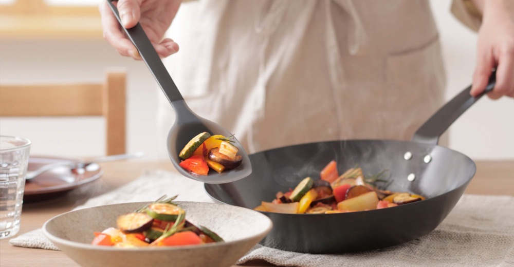 烹飪中，一手持長柄矽膠料理匙盛裝熱騰騰的炒蔬菜，另一手握著煎鍋，鍋中也盛有相同的炒蔬菜，鍋中冒著熱氣，旁有一碗裝著蔬菜，桌上還有水杯和餐具。