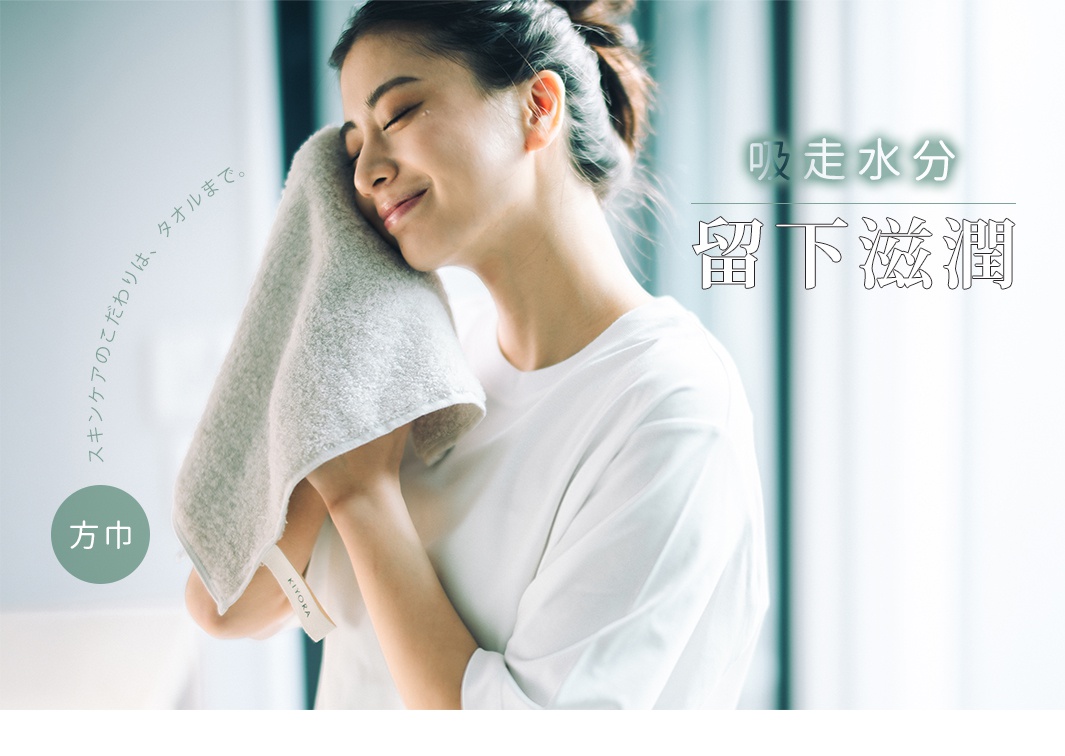 一位留著低馬尾的女性正閉眼享受地用灰色柔軟方巾輕撫臉龐，方巾邊緣繡有 KIYORA 字樣，畫面右側標題寫著吸走水分留下滋潤，左側則有強調護膚細節連毛巾都要講究的文字，整體氛圍溫馨且具居家質感。