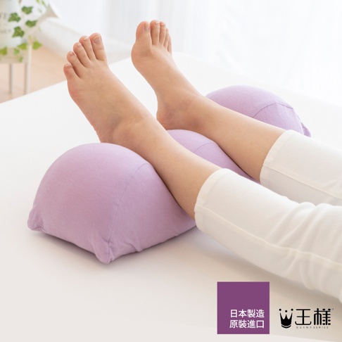淺紫色布面抬腿枕，分為兩段式設計，支撐女性雙腿，露出腳部，背景為白色床鋪。