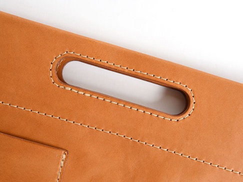 職人手作皮革橫式收納袋中袋，以淺棕色皮革製成，車縫線為白色。圖片特寫了袋子上的提把，提把開口處有精緻的縫線加固，袋身有多條平行車縫線，展現皮革的紋理與質感。