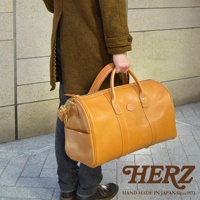 職人手作皮革拉鍊波士頓包，採用天然皮革製成，呈現溫潤的焦糖棕色。包身線條簡潔，配有雙提把與側邊插袋，適合肩背或手提。精緻縫線展現細膩工藝。