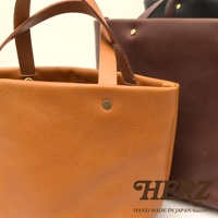 職人手作皮革托特包，前方為焦糖棕色，後方為深棕色，均為柔軟皮革材質，配有同色系皮革提把與金色金屬釦。