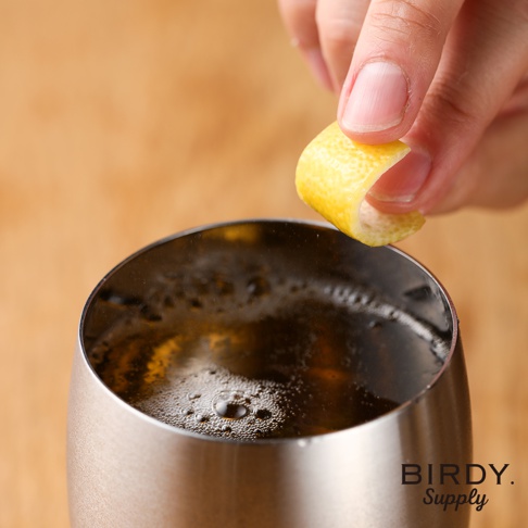 不鏽鋼啤酒杯中裝有深色飲料，表面佈滿細小氣泡。一隻手正將檸檬皮捲插入杯中，杯身為光滑的銀色不鏽鋼材質。