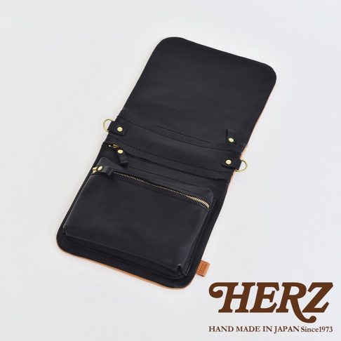 職人手作皮革翻轉雙色直式斜背包，主體為黑色皮革，翻蓋內側及斜背包外層下方為原色皮革，設有拉鍊小袋及金屬掛環。