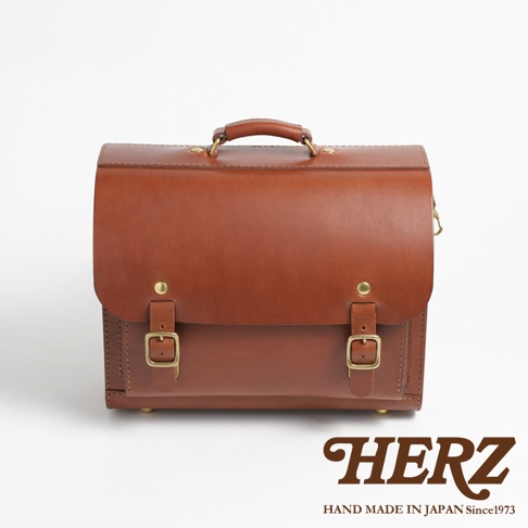 職人手作皮革橫式書包，棕色皮革，配有金屬扣環和提把，底部有四個金屬腳釘。