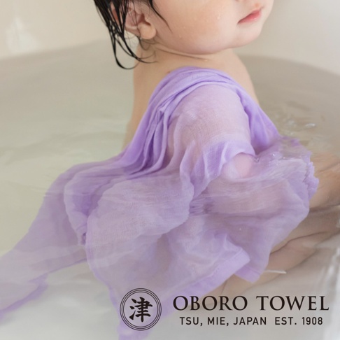 嬰兒頭部特寫，懷中抱著一條淡紫色三層紗純棉裹身毯，毯子浸在水中，部分濕潤。