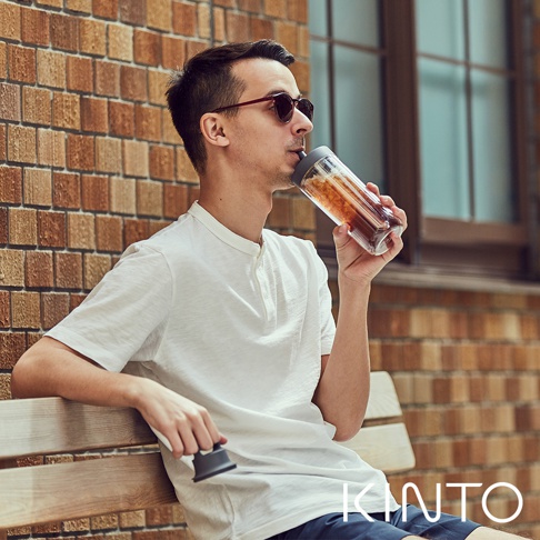 男子手持TO GO BOTTLE雙層隨手瓶，瓶身透明，內有茶色飲品，瓶蓋為深灰色，搭配白色短袖上衣和藍色短褲，坐在木質長凳上。
