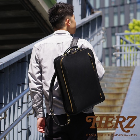 職人手作黑色皮革商務電腦後背包 R-25，搭配金色拉鍊，一個男性背著走在樓梯上。