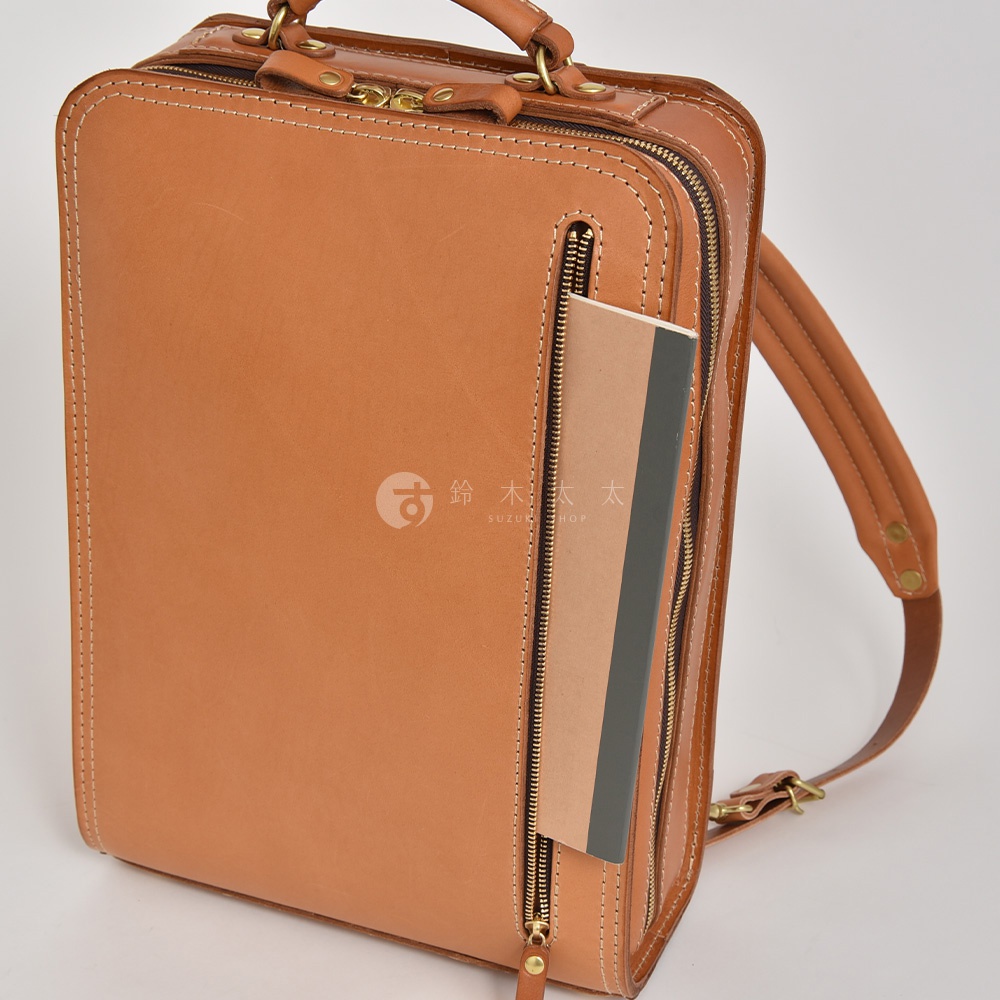 一件棕褐色皮革製成的直立式後背包，配有拉鍊開合、頂部提把及可調節肩帶。背包正面有一個垂直拉鍊口袋，內含一本筆記本。