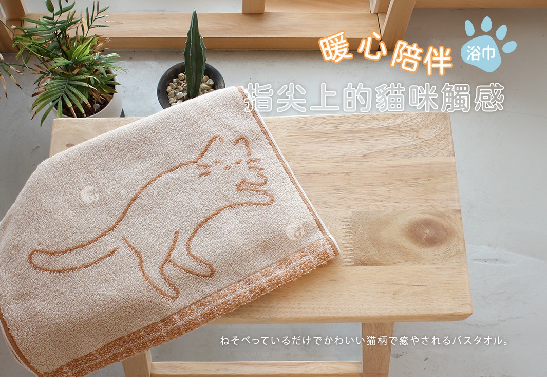 這是一條放在淺色木質小凳上的淺棕色浴巾，浴巾表面以簡約線條勾勒出貓咪伸懶腰的圖案，整體呈現溫暖舒適的氛圍，背景上方點綴著關於療癒貓咪觸感的日文與中文說明文字。