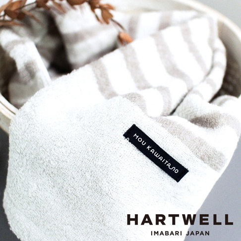 今治毛巾，白色搭配淺棕色條紋，材質柔軟，繡有「MOU KAWAITA」字樣的黑色標籤，上方有乾燥的裝飾性枝葉。