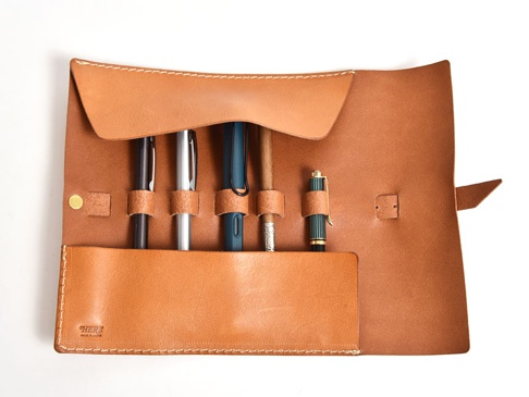 職人手作淺棕色皮革捲捲收納筆袋，內有五個筆槽，分別裝有黑色、銀色、藍色、木質和綠色鋼筆。筆袋以白色縫線裝飾，並以黃銅色鈕扣和皮革帶固定。