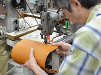 一位師傅正使用工業縫紉機，縫製一個淺棕色皮革製成的 A4 筆記本書套。皮革表面光滑，邊緣有縫線。