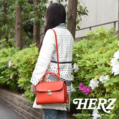 日本HERZ職人手作紅色皮革多功能收納單肩包，模特兒側身站立於綠色植栽旁，展現包款手提、肩背三用設計