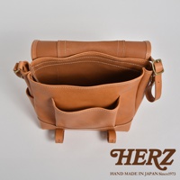 tan皮革復古郵差包，有縫線裝飾，前方有兩個小口袋，側邊有綁帶孔，上方有翻蓋及金屬釦具。