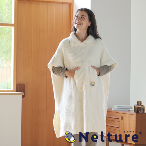 一位年輕女子穿著一件米白色的羊羔絨斗篷，斗篷上有個大口袋，上面還縫著小標籤，她雙手插在口袋裡，臉上帶著微笑看向遠方。