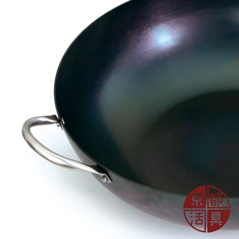 一個深色的圓形鐵鍋，鍋身呈深藍色帶有金屬光澤，左側有一個銀色的弧形提把。