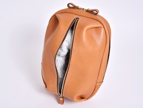 淺棕色皮革製成的立體斜背包，拉鍊部分敞開，露出內部淺灰色毛巾狀物品，搭配金色拉鍊和皮革拉環，縫線明顯。