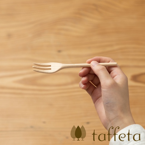 一隻手拿著一支淺色楓木製成的點心叉，叉子有三齒，背景是木紋桌面。