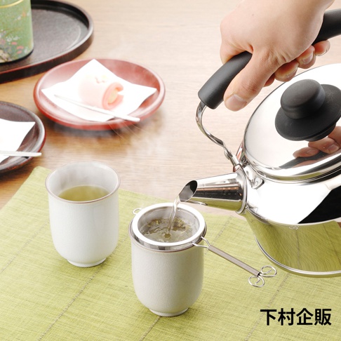 不鏽鋼深型濾茶網，濾網網眼細緻，帶有長柄，置於白色陶瓷茶杯中，旁有一杯綠茶，上方為手持銀色不鏽鋼茶壺，壺嘴正將熱水注入濾網。