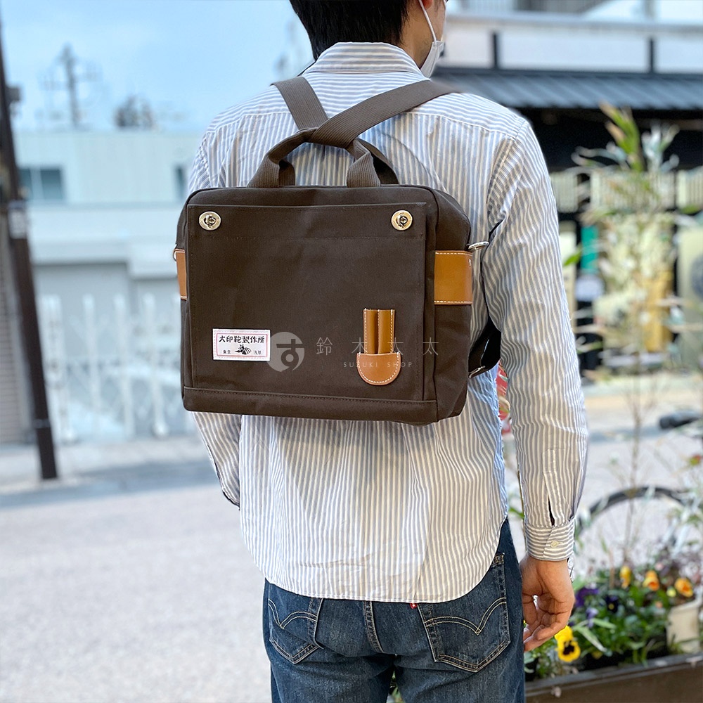 深棕色純棉帆布製成的三用側背包，配有棕色皮革飾邊和提把，背包帶為灰色。