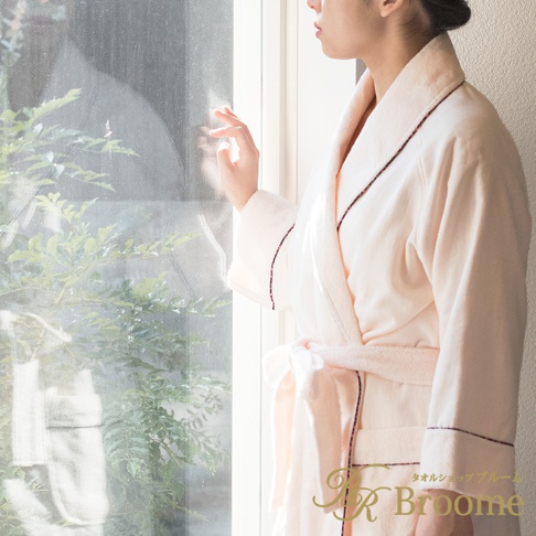 身著淺粉色今治純棉浴袍的女性，浴袍有深色滾邊，腰間繫帶，面向窗戶。