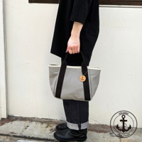 灰色帆布製手提托特包，配有黑色提把和白色內襯，包包上掛有船錨造型的木質吊飾。