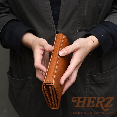 一名穿著深灰色外套的人雙手捧著一只棕色皮革長夾，長夾呈現手風琴式的多層風琴褶設計，側面顯示出細緻的車縫線與厚實的真皮質感，右下角印有HERZ品牌標誌與手工製作說明文字。