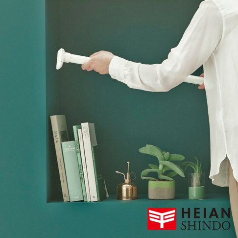 白色伸縮置物架由手持，兩端有圓形底座，置於深綠色牆面內嵌式層架上，層架上擺放幾本淺綠色書本、金色噴霧瓶及盆栽。