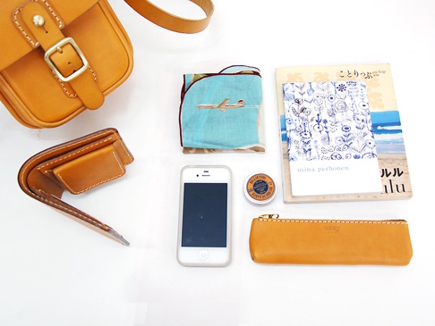 職人手作皮革皮扣直式單肩包 (CP-4)，搭配同色系皮夾、手機、小圓罐、印有飛機圖案的淺藍色手帕，以及一本藍色花紋封面書籍。