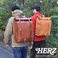 兩款職人手作皮革後背包，分別為深棕色與淺棕色，皆採用捲口設計，配有皮質揹帶、金屬釦具及側袋，適合戶外活動使用。