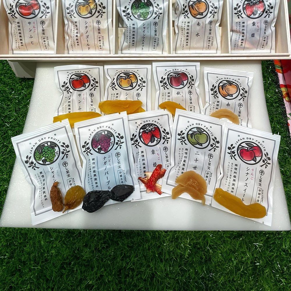 長野縣南信州果乾20入桐木禮盒，展示多種口味的獨立包裝果乾，包裝袋為白色，印有水果圖案與日文品名，包含葡萄、草莓、蘋果、柿子等，果乾顏色有黃色、紫色、紅色、褐色等，部分已從包裝中取出，堆疊於白色底座上。