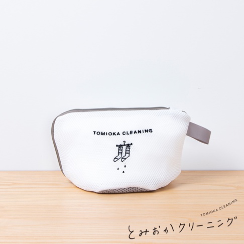 白色網狀材質的貼身衣物洗衣袋，配有灰色滾邊和提把。袋面印有黑色「TOMIOKA CLEANING」字樣及一對掛在衣架上、滴水的襪子圖案。