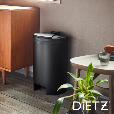 黑色圓柱形自動感應靜音垃圾桶，側面有開關按鈕，頂部有斜面開蓋設計，材質為霧面塑膠，置於木質地板旁，與木櫃和茶几一同入鏡。
