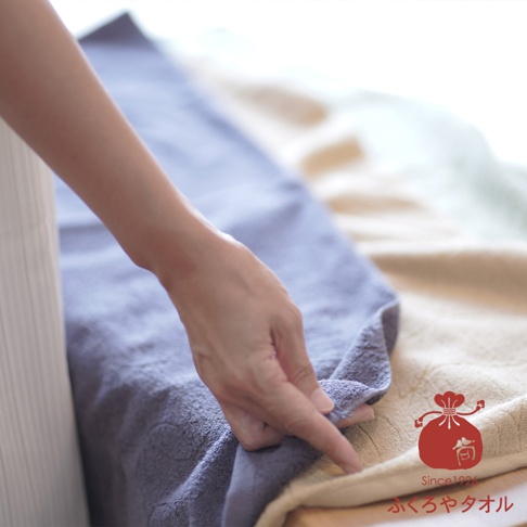 一隻手正觸碰著一條藍紫色的毛巾，其下方疊著一條米色的毛巾，背景還有淺綠色的毛巾。毛巾材質看起來柔軟，帶有細緻的紋理。