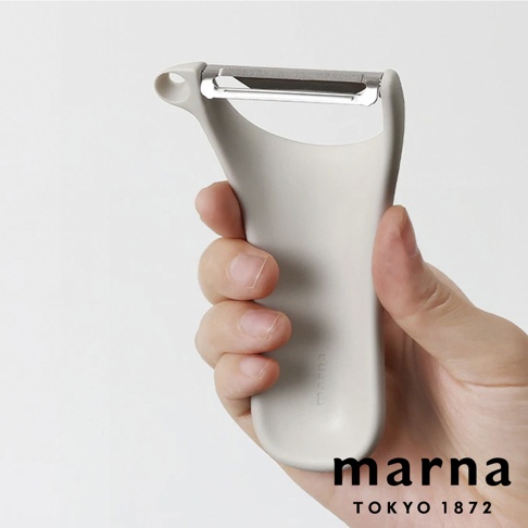 一隻手握著淺灰色簡約風格的 Marna 品牌削皮器，該削皮器擁有符合人體工學的圓潤握把設計，上方配有不鏽鋼刀片與圓孔吊掛設計，整體造型流暢簡潔。