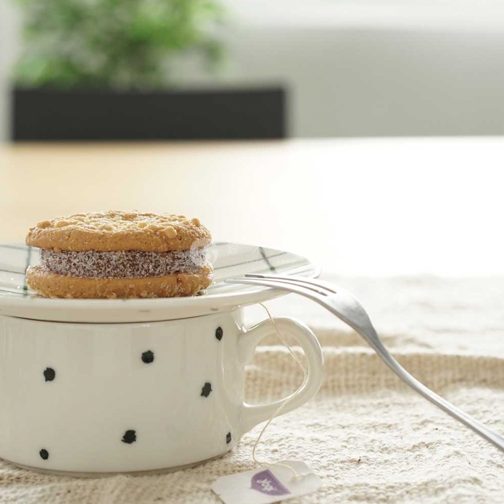 一個白色陶瓷手繪馬克杯，杯身點綴著黑色圓點。杯蓋上放著一個夾心餅乾，旁邊有一支叉子和一個茶包。