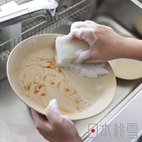 一隻手正拿著沾滿泡沫的洗碗海綿清洗一個有番茄醬污漬的淺色盤子，盤子放在水槽裡，背景有餐具瀝水架。