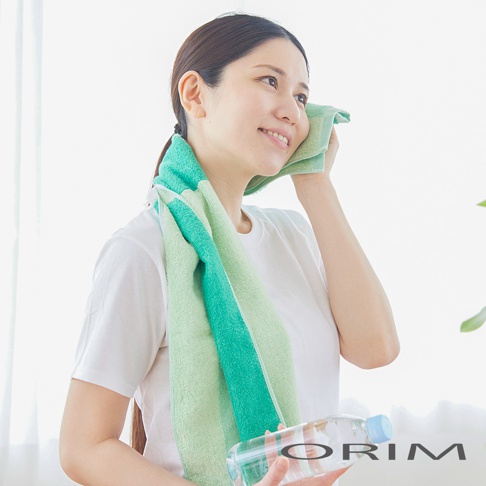 一位女子使用一條綠色和淺綠色拼接的毛巾擦拭臉部，毛巾掛在脖子上。她手裡拿著一瓶水。