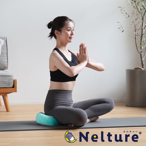 一位女性盤腿坐在瑜珈墊上，雙手合十於胸前，身穿黑色運動內衣和灰色瑜珈褲，坐墊為土耳其藍色，背景為簡潔的室內空間，右側有盆栽植物。