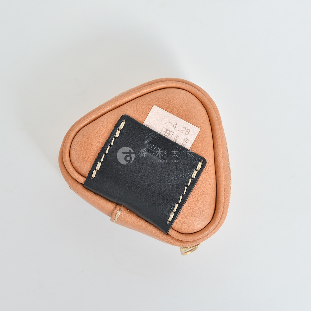 職人手作皮革三角飯糰造型小物收納包，主體為淺棕色皮革，頂部有一塊方形黑色皮革裝飾，以白色縫線車縫。收納包上有一張標籤。