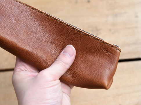 手持一只棕色皮革製成的Organ三角收納筆袋，表面紋理清晰，帶有拉鍊。