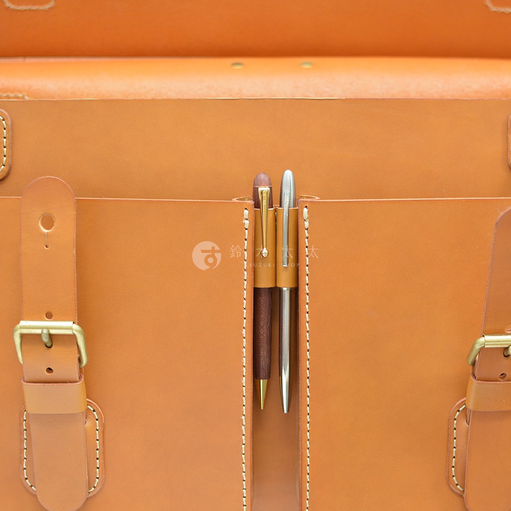 職人手作皮革旅行包 A-102，特寫展示包身正面，包含兩個筆插，分別插入一支深棕色筆和一支銀色筆。包身採用天然皮革，顏色為暖棕色，縫線清晰。左側有皮帶扣設計。