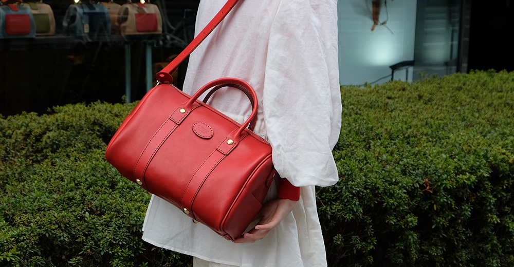 紅色皮革波士頓包，配有可拆卸肩帶和雙提把，包身有拉鍊開合，底部有四個金屬腳釘，整體為簡約的圓筒形設計。