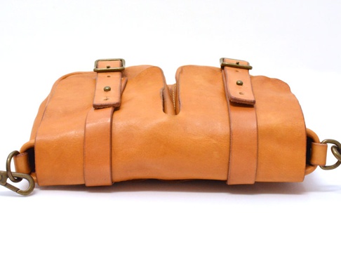 職人手作皮革水洗加工前雙口袋肩背包，呈現溫暖的棕褐色調。包款採用天然皮革材質，表面帶有細緻紋理。中間有明顯的開口設計，兩側各有一個皮帶扣飾，並配有金屬鉤扣的肩帶。