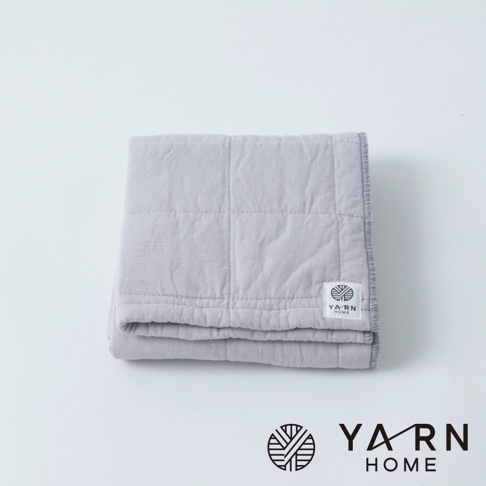 一條淺灰色、表面有格紋縫線的毛巾，材質為脫脂棉混紗，邊緣有細緻的車縫線，角落處有一個印有「YARN HOME」字樣的白色標籤。