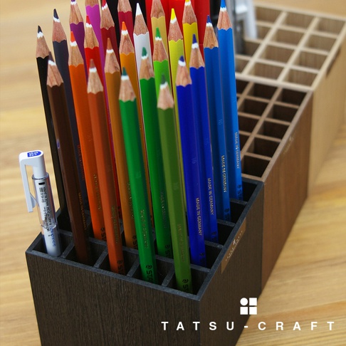 深棕色木質筆筒，內含多支不同顏色的鉛筆，如紅色、橘色、綠色、藍色等，部分鉛筆上有「MADE IN GERMANY」字樣。筆筒旁有一支白色筆。