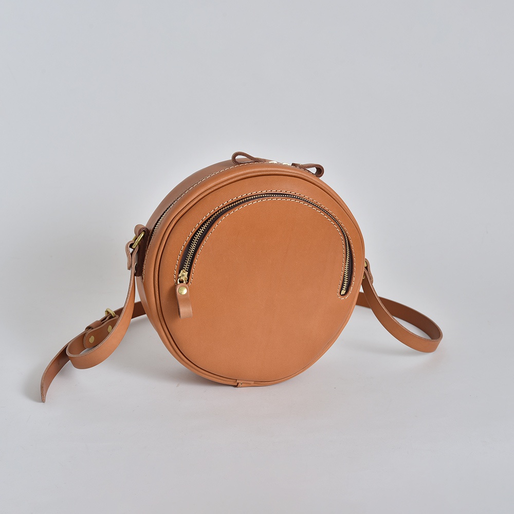 職人手作皮革小圓包，呈現自然棕色調，採用真皮材質，圓潤造型，配有兩條拉鍊開合，金屬拉頭，細緻縫線，附可調節長度的斜背帶。