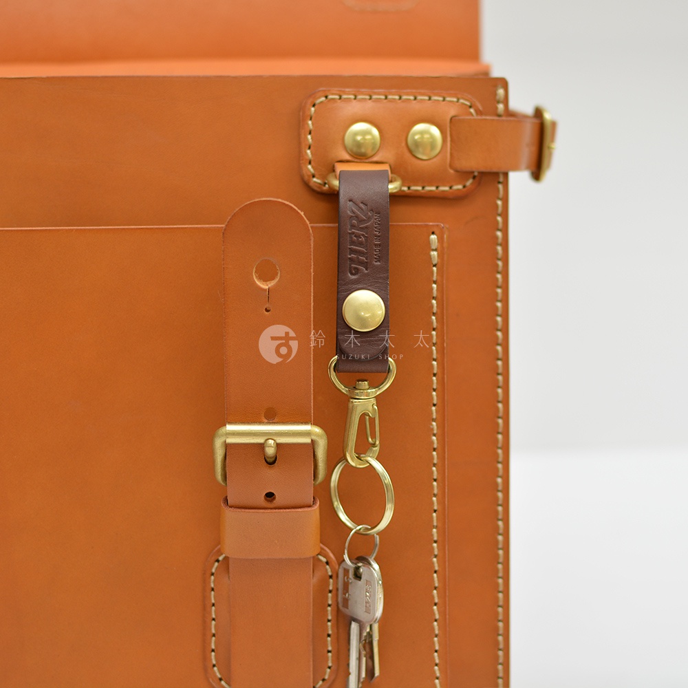 職人手作皮革旅行包A-102，展示的是包包的局部特寫，以溫暖的棕色皮革為主體，搭配金屬釦環和鑰匙串。皮革表面有細緻的縫線，展現職人手工的質感。